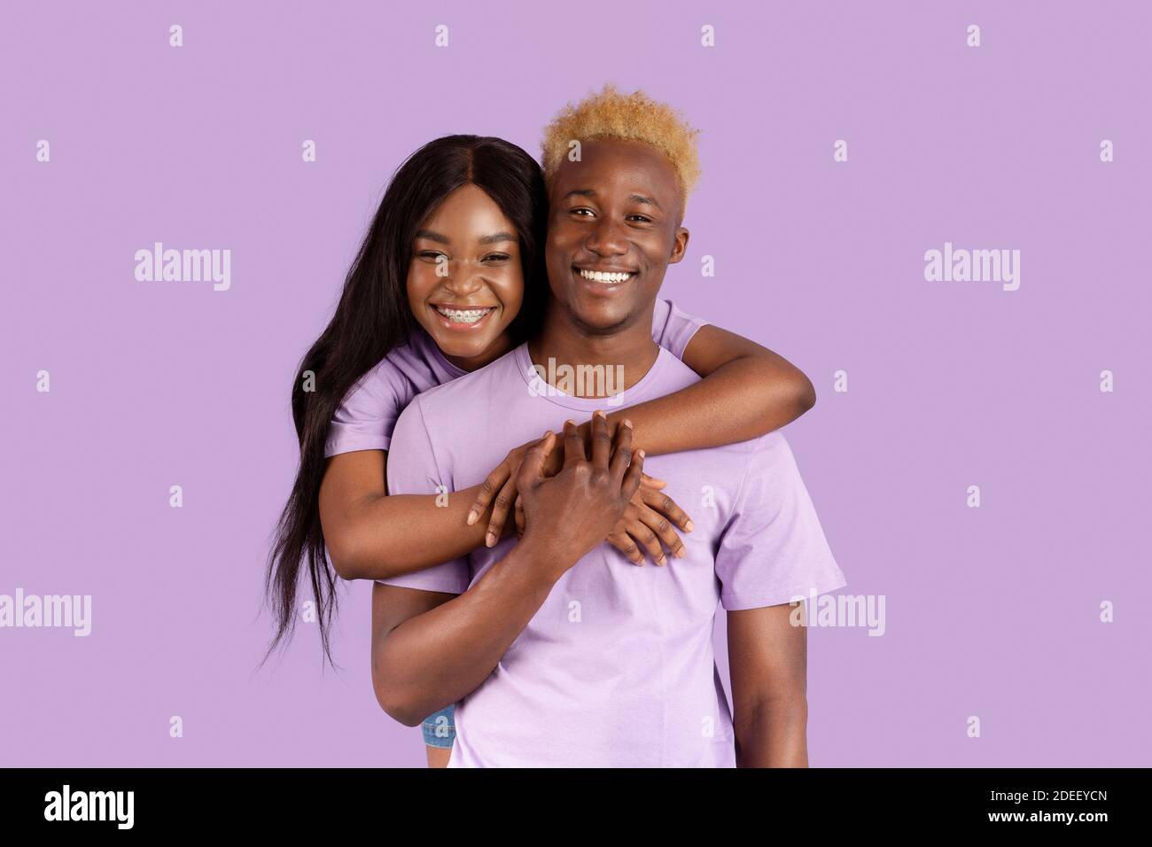 Hermosa mujer negra abrazando a su novio desde atrás, sintiéndose enamorada y feliz sobre fondo de estudio lila Foto de stock