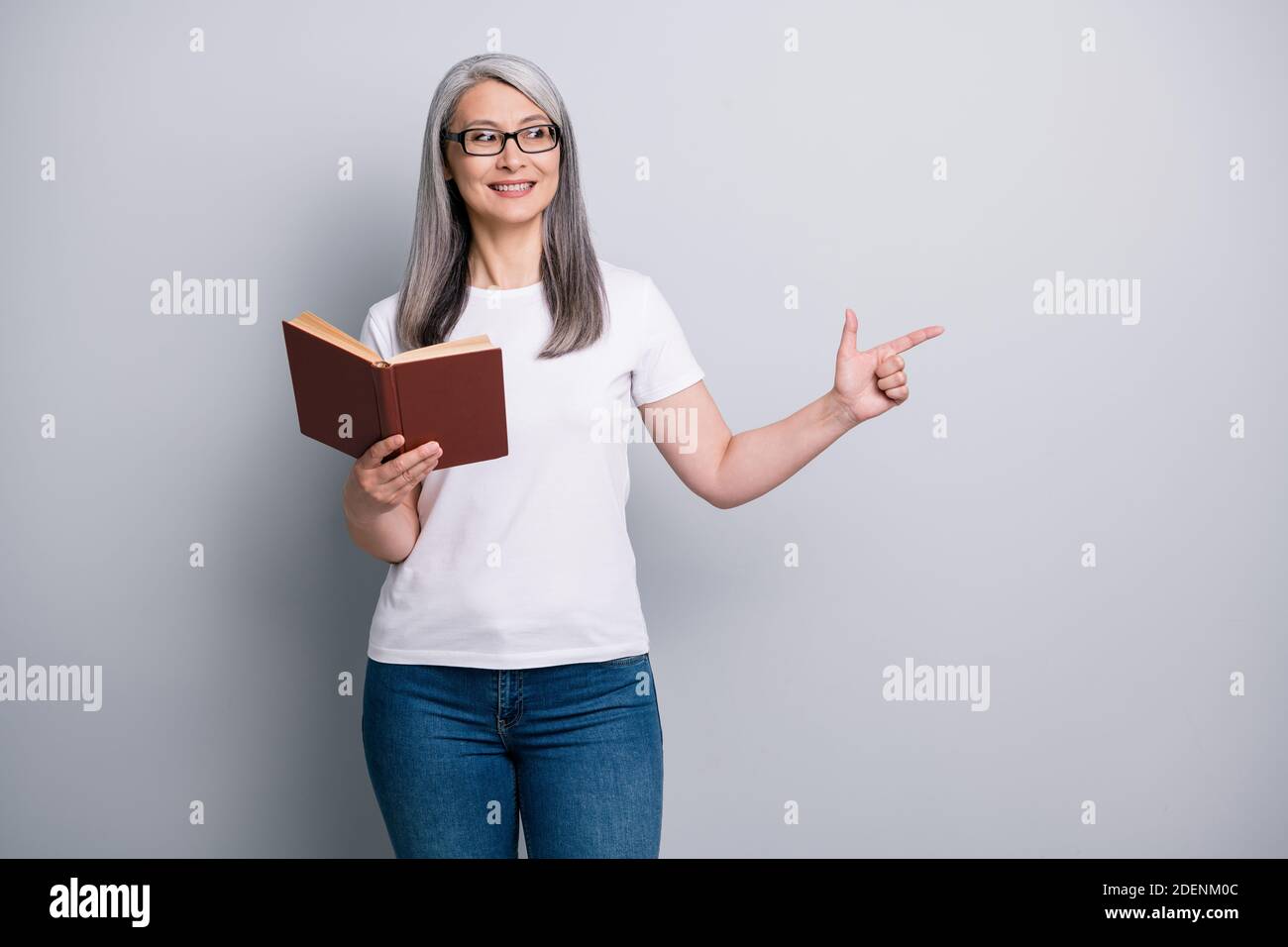Foto de adorable señora mayor informal conjunto gafas sujeción libro mirando dedo apuntador espacio vacío gris Fotografía de stock - Alamy
