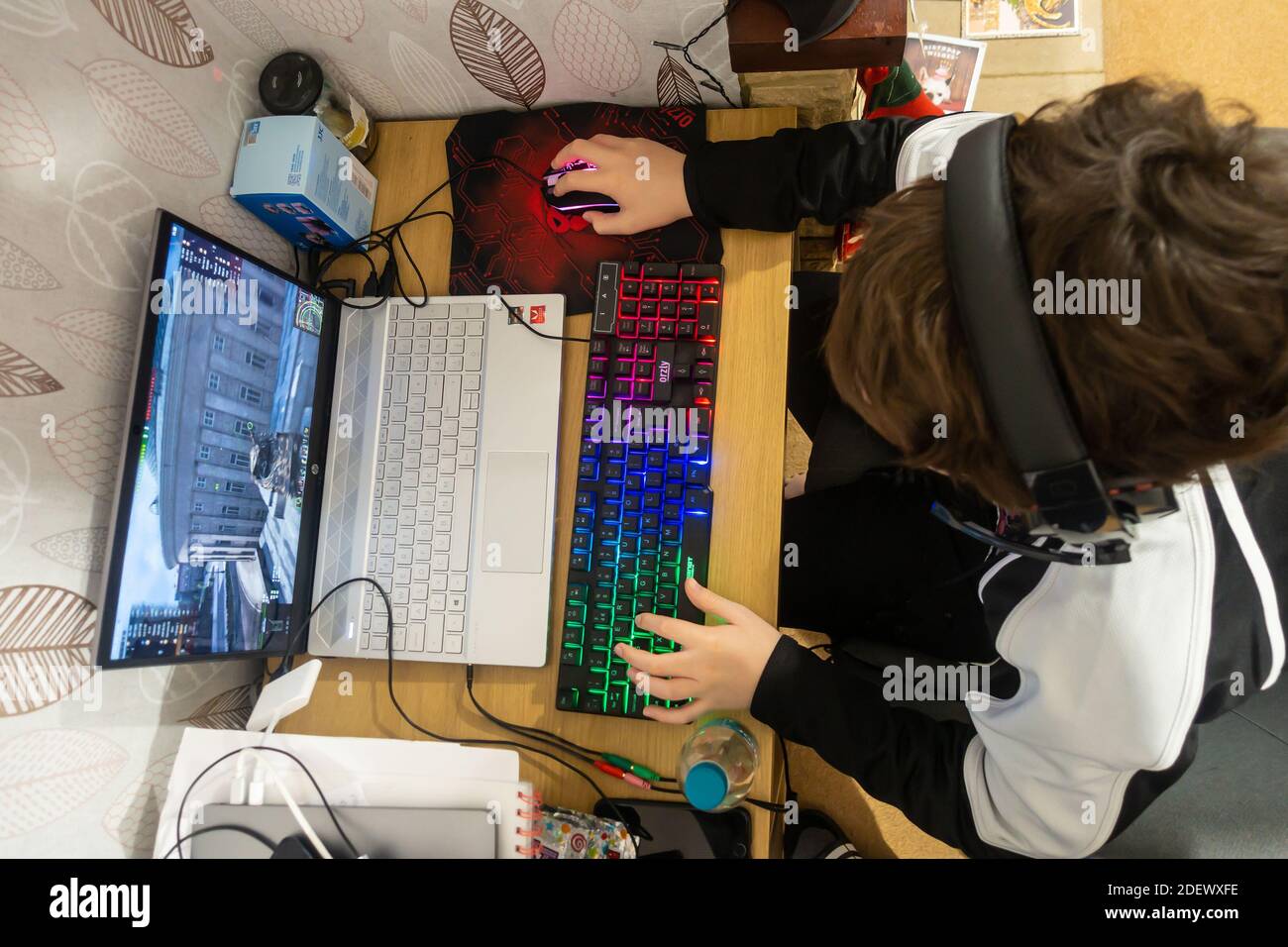Mirando hacia abajo a un niño sentado en un escritorio, jugando un juego de ordenador en un ordenador portátil con un ratón y un teclado de juego iluminado. Foto de stock