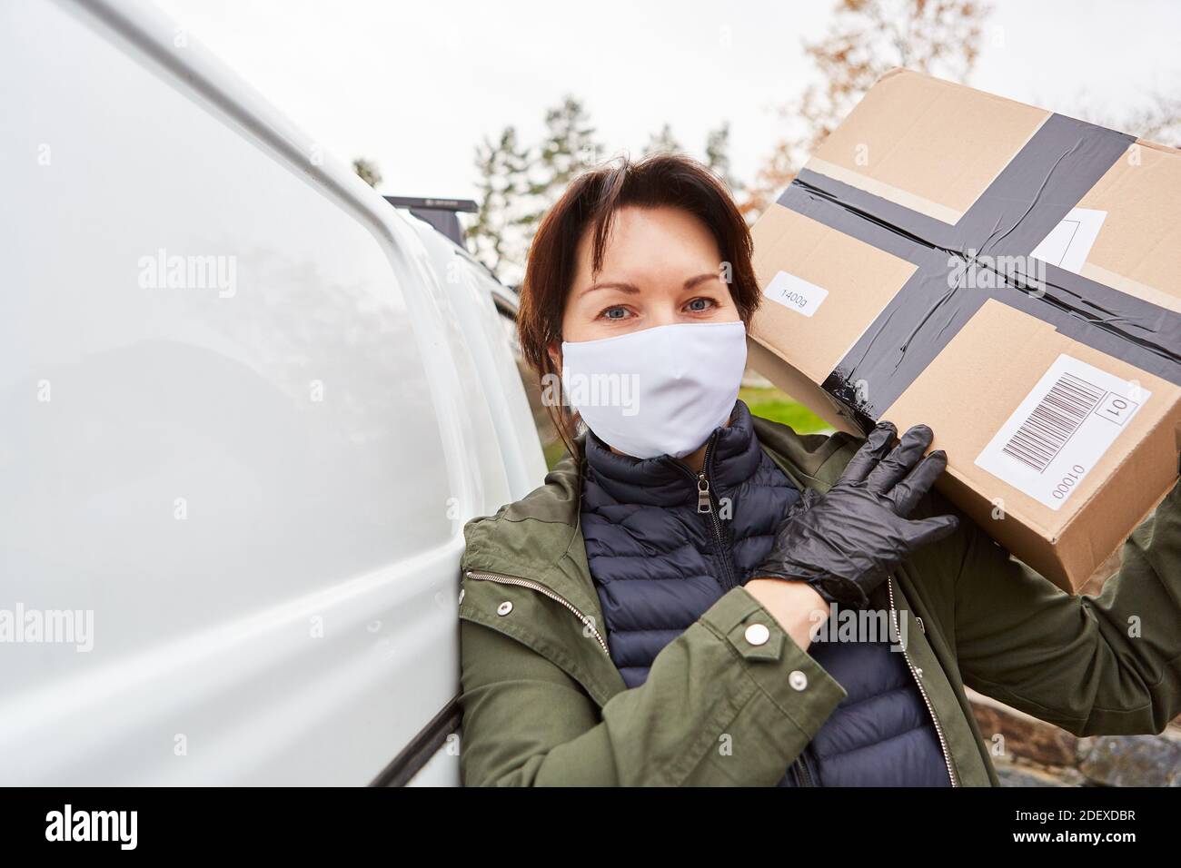 El transportista de entrega de paquetes con máscara frontal lleva el paquete de la furgoneta para la entrega Foto de stock