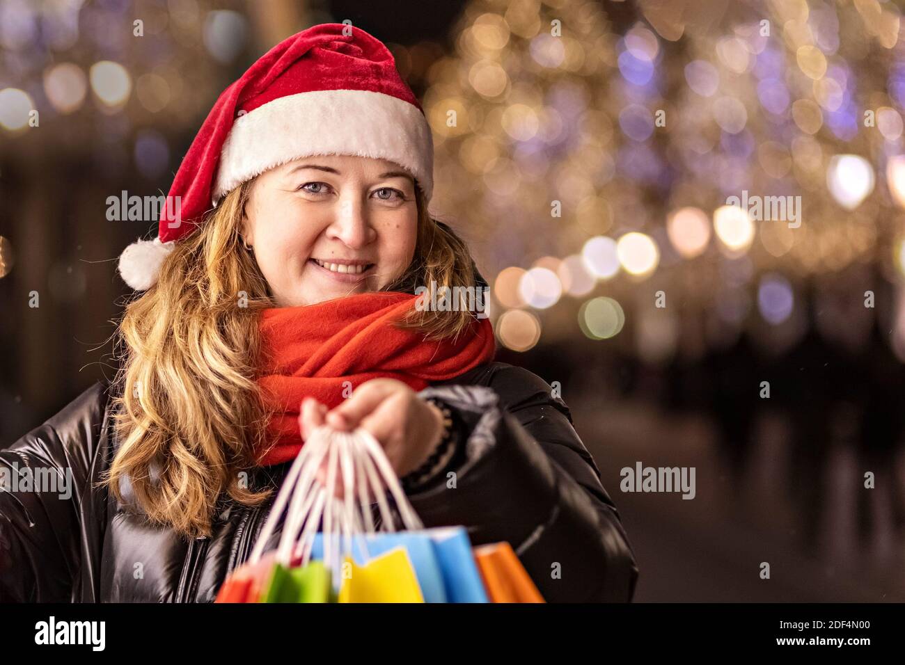 Una mujer con pelo largo y un sombrero de Santa cerca de la ventana de una tienda de la ciudad con compras en bolsas de papel de colores. Compras de año Nuevo. Foto de stock