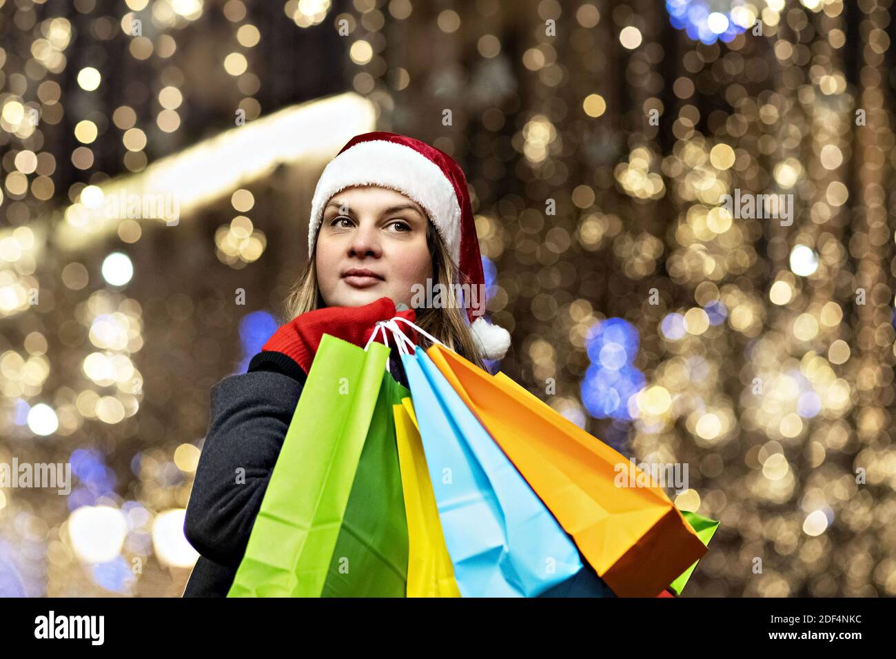 Una mujer con pelo largo y un sombrero de Santa cerca de la ventana de una tienda de la ciudad con compras en bolsas de papel de colores. Compras de año Nuevo. Foto de stock