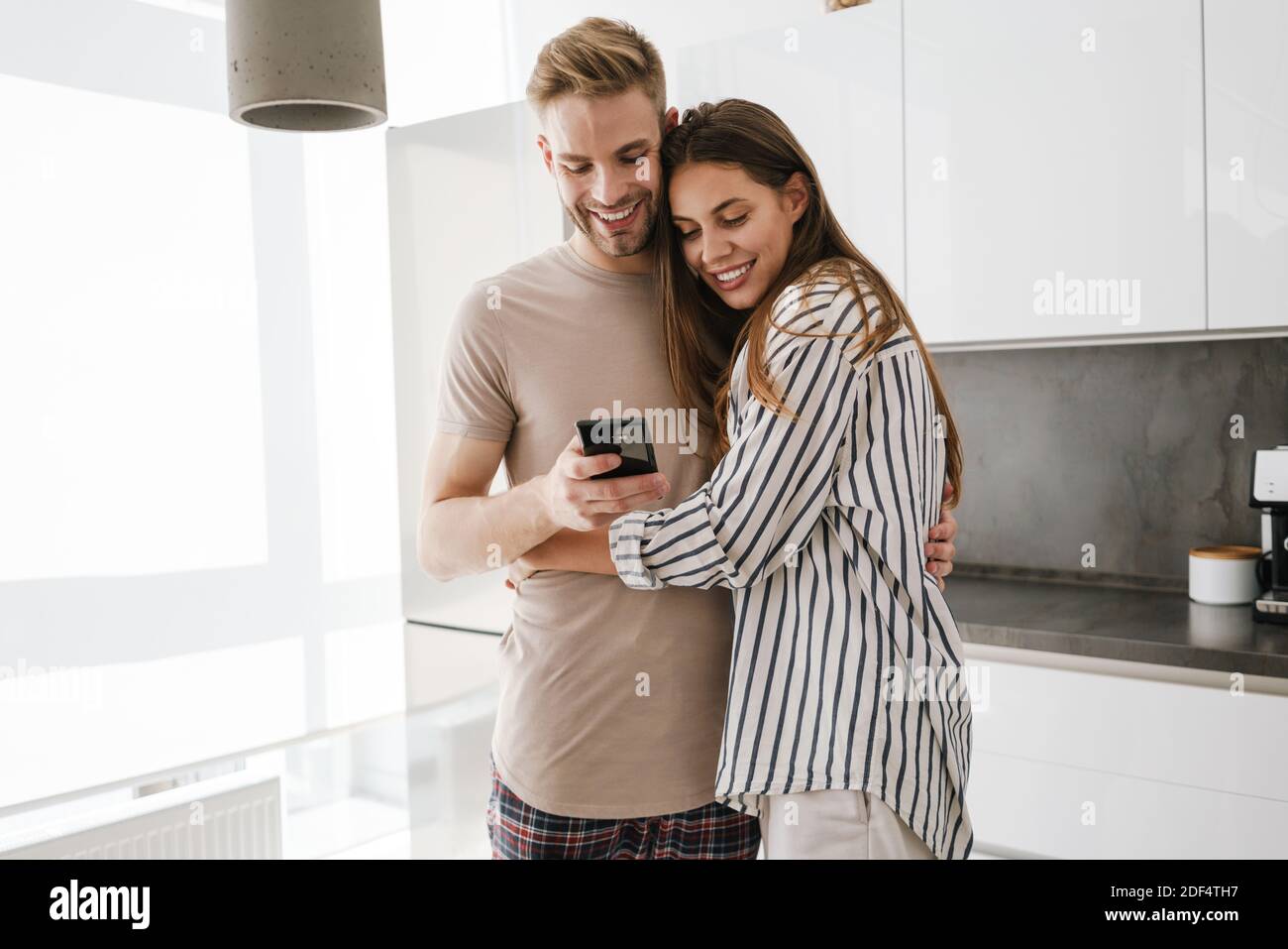 Joven hermosa pareja caucásica en ropa de ocio abrazando y sosteniendo teléfono móvil en casa Foto de stock
