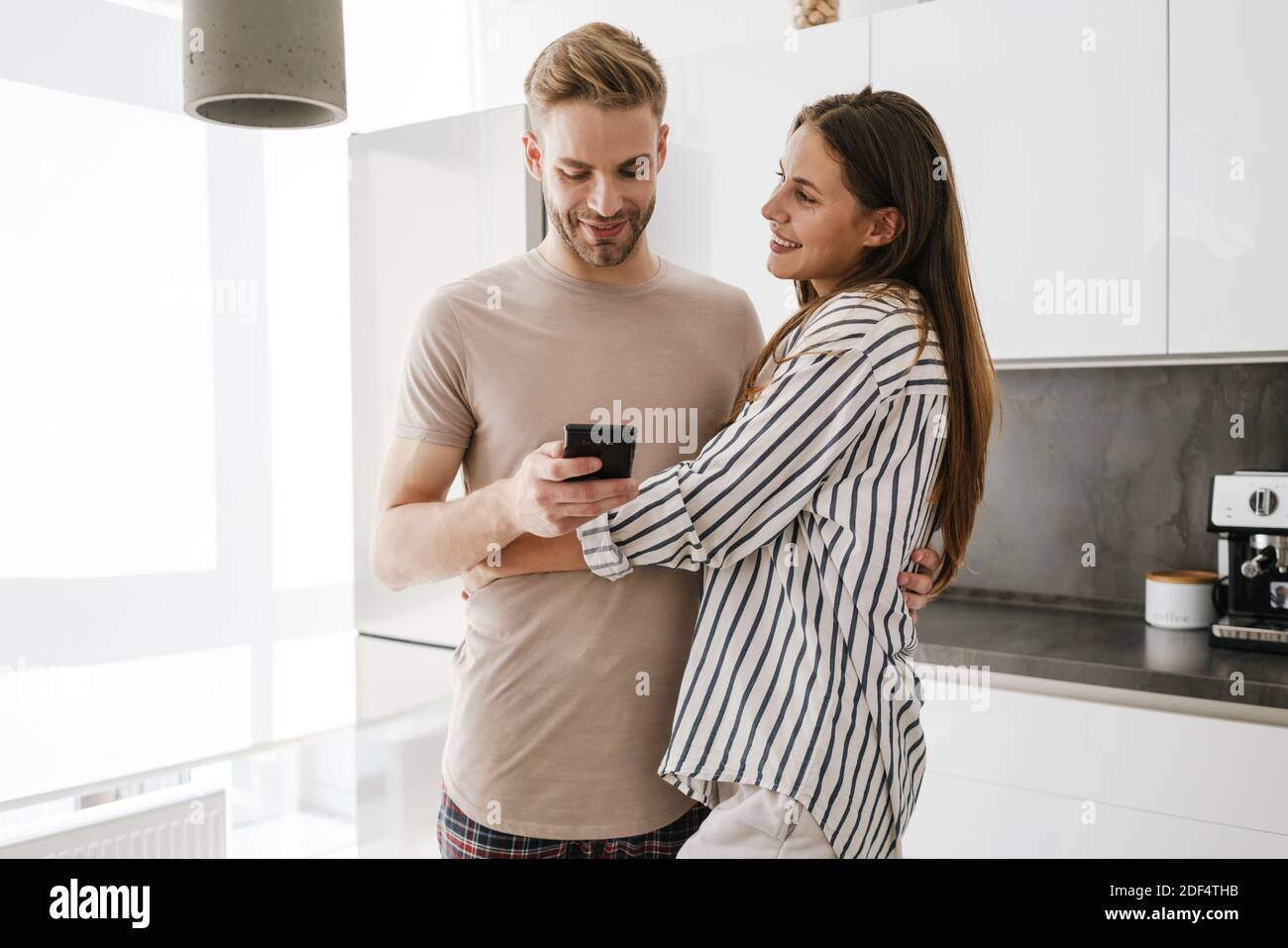 Joven hermosa pareja caucásica en ropa de ocio abrazando y sosteniendo teléfono móvil en casa Foto de stock