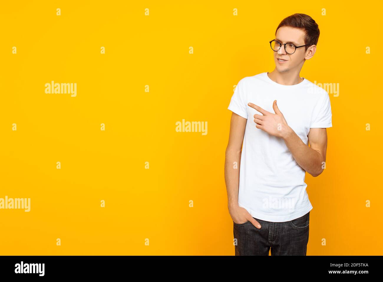 Hombre atractivo, con gafas, en blanco camiseta, apuntando con dos dedos al lado, apuntando al espacio vacío, sobre fondo amarillo Foto de stock
