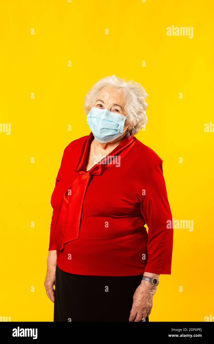 Retrato de estudio de una mujer mayor con camisa roja y máscara facial quirúrgica, sobre un fondo amarillo Foto de stock