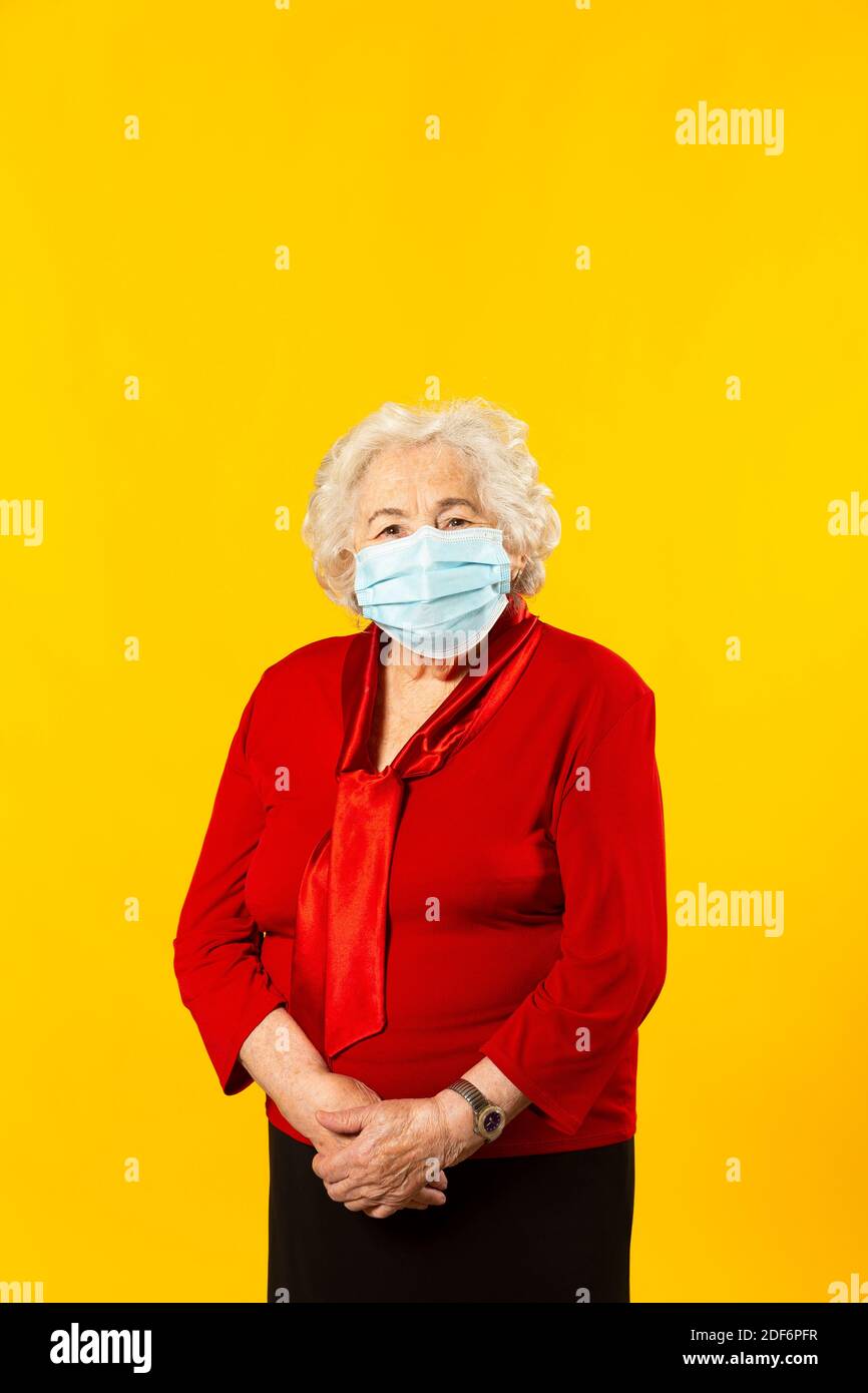 Retrato de estudio de una mujer mayor con camisa roja y máscara facial quirúrgica, sobre un fondo amarillo Foto de stock