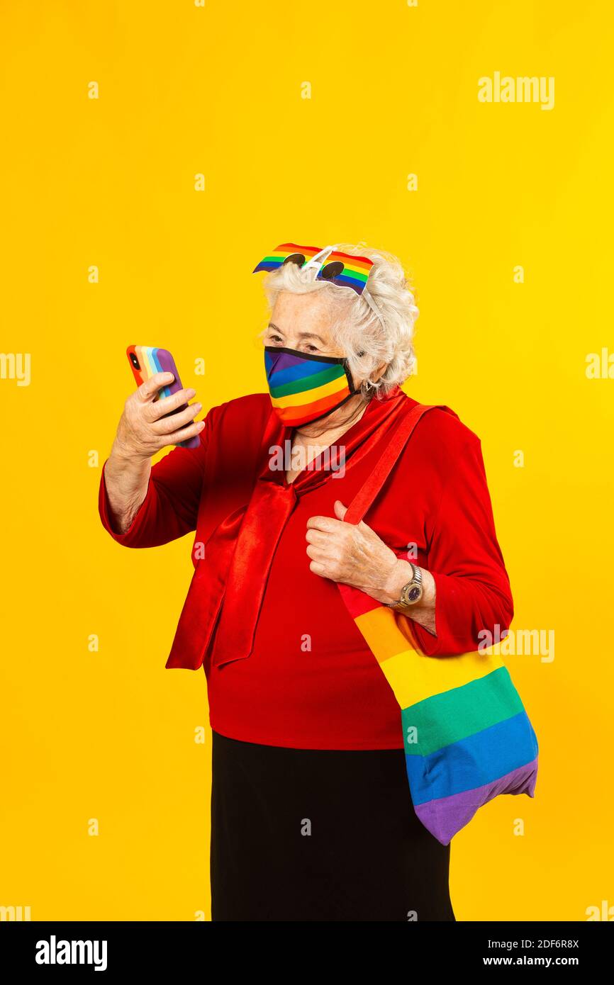 Retrato de estudio de una mujer mayor con camisa roja, gafas de sol arcoiris, máscara facial de color arco iris y una bolsa, teniendo una videollamada con su móvil Foto de stock