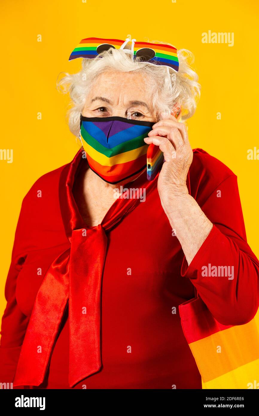 Retrato de estudio de una mujer mayor con camisa roja, gafas de sol arcoiris, máscara facial de color arco iris y una bolsa, teniendo una llamada con su teléfono móvil, Foto de stock