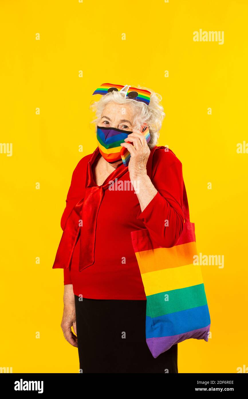 Retrato de estudio de una mujer mayor con camisa roja, gafas de sol arcoiris, máscara facial de color arco iris y una bolsa, teniendo una llamada con su teléfono móvil, Foto de stock