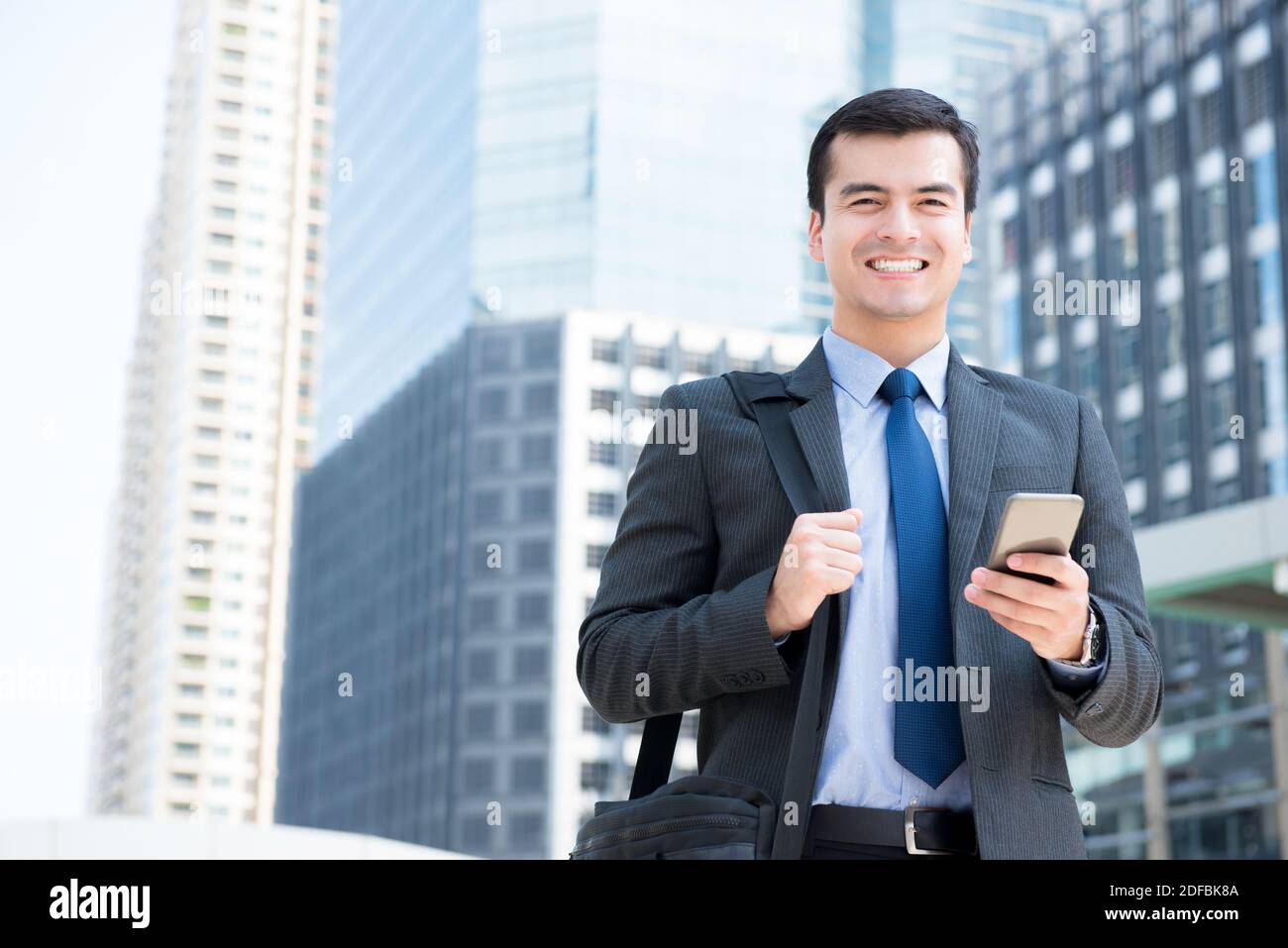 Un hombre de negocios sonriente usando el teléfono móvil mientras llevaba la bolsa en un borroso fondo del edificio de la ciudad - viajes de negocios y conceptos de roaming móvil Foto de stock