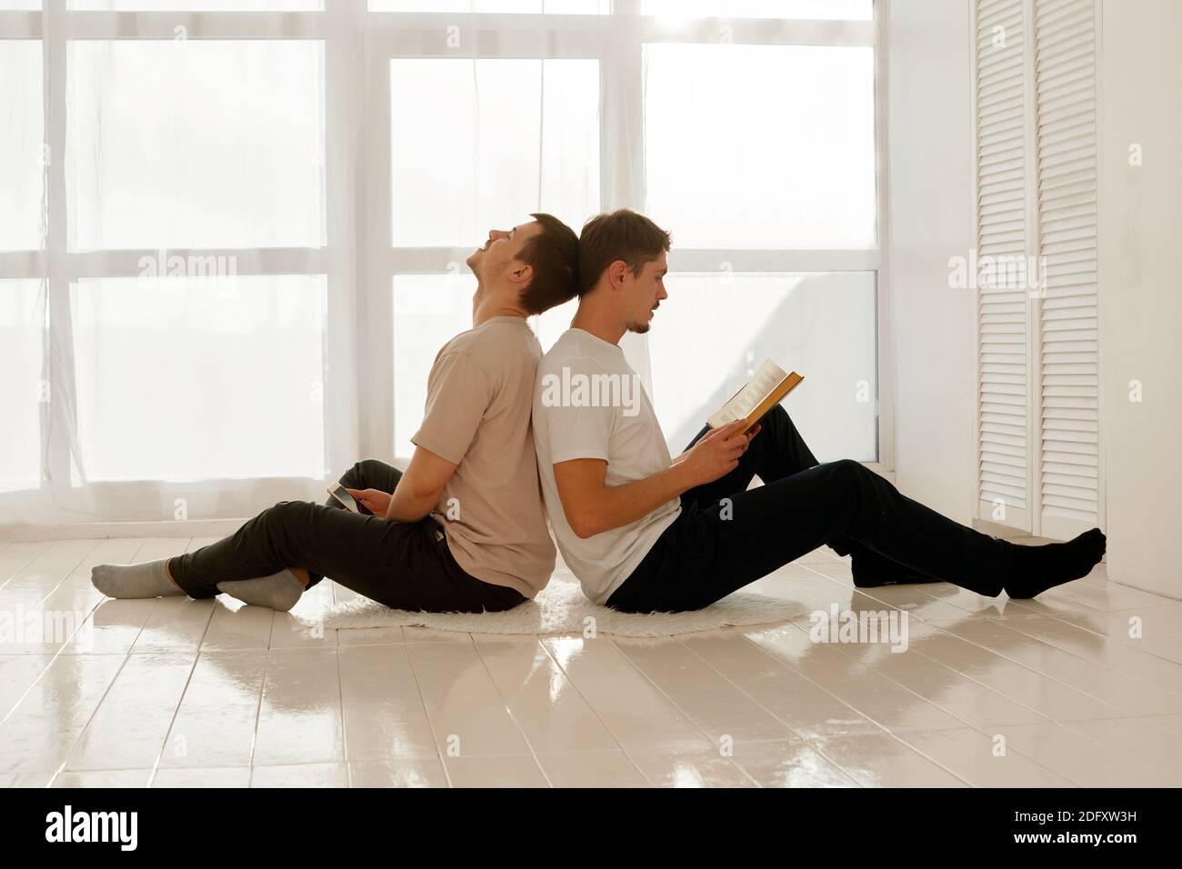 Un chico agradable lee un libro y su amigo lo escucha y mira en un smartphone. Un par de hombres gays pasan tiempo en casa sentados en el piso de la ho Foto de stock