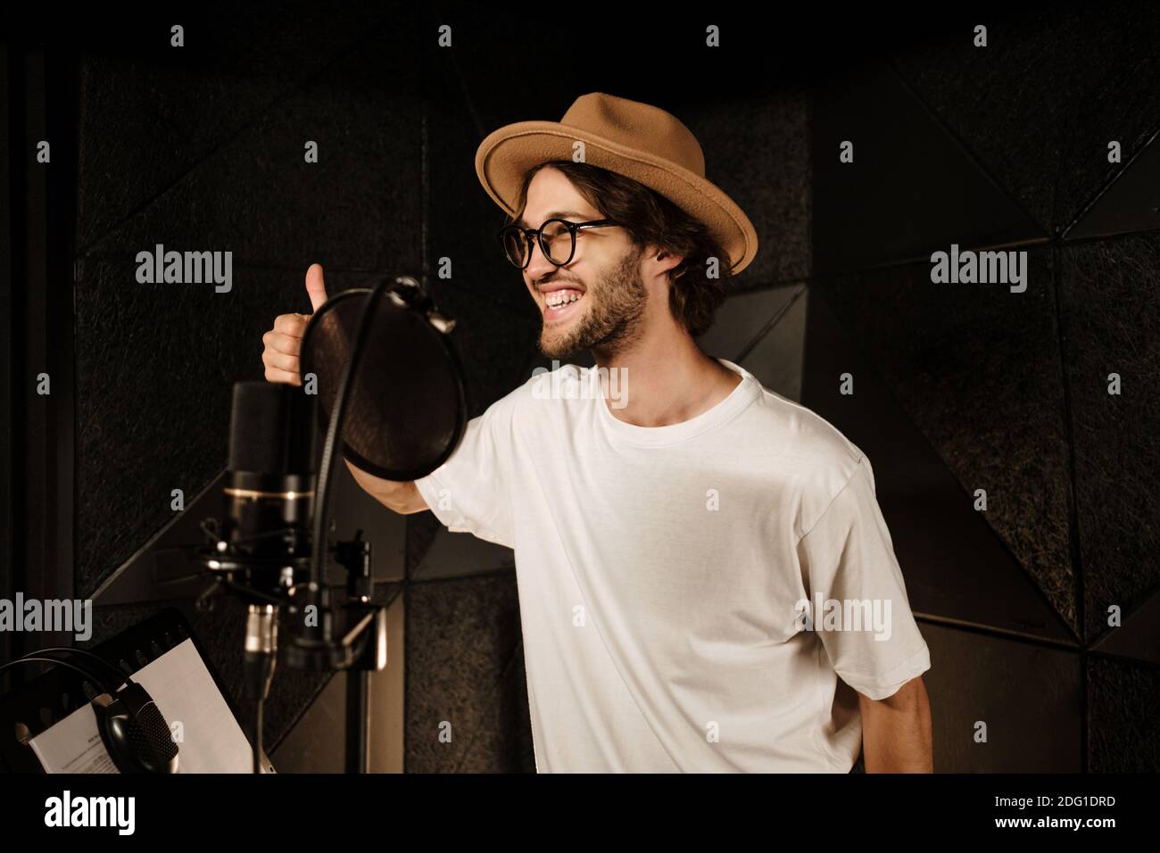 Atractivo y elegante cantante masculino felizmente mostrando el pulgar hacia arriba grabando nueva canción en estudio moderno. Como gesto Foto de stock