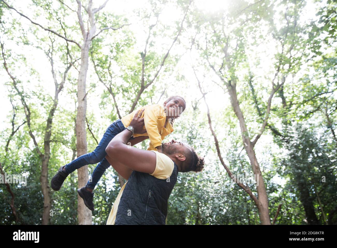 Juguetón padre sin preocupaciones levantando a hija debajo de los árboles en el parque soleado Foto de stock
