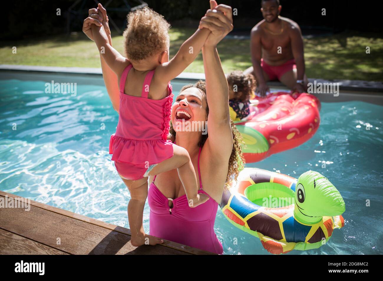 Madre juguetona levantando a su hija pequeña en la piscina soleada Foto de stock
