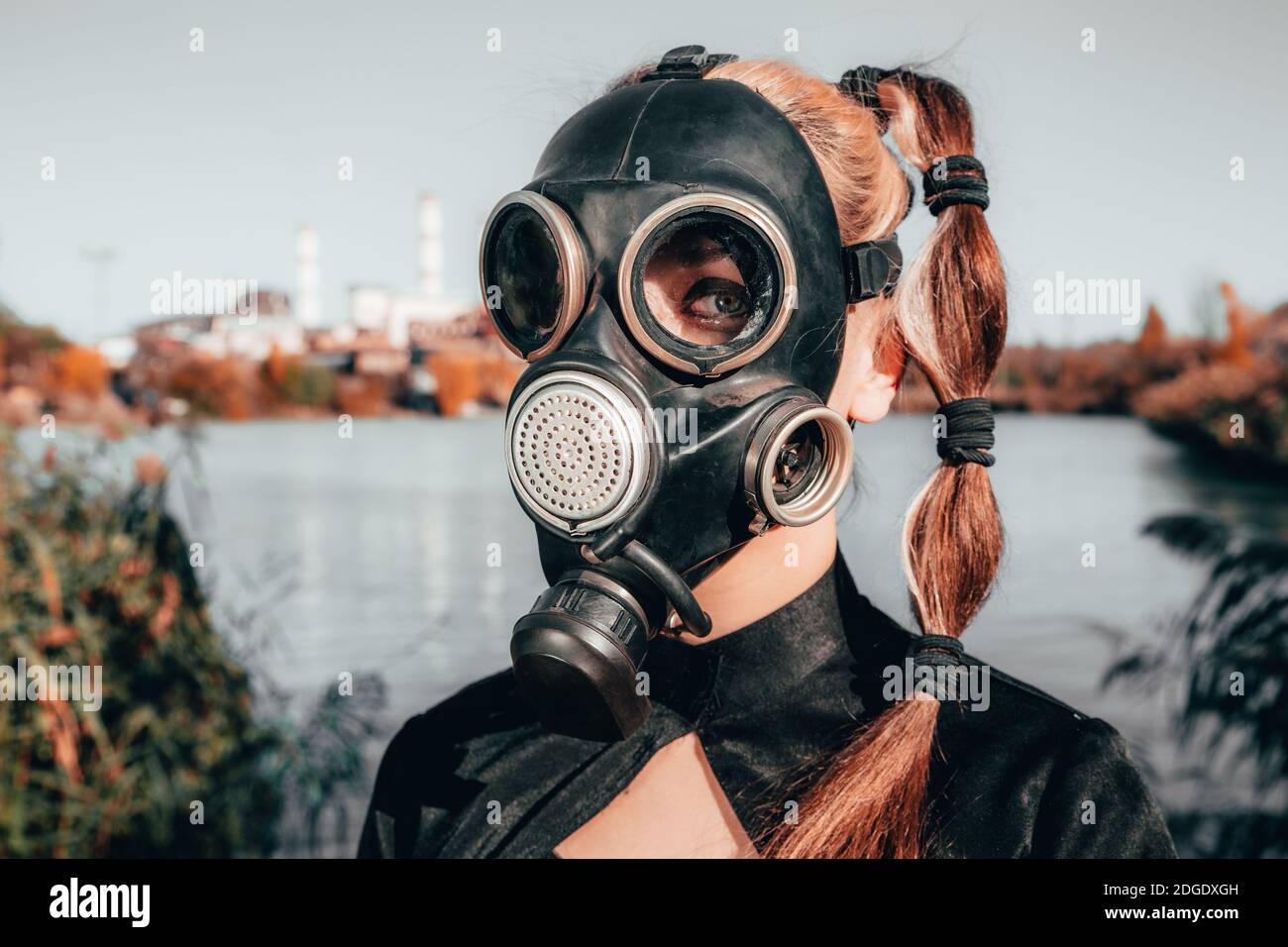 niña en una máscara de gas en un vestido rojo con un libro en sus manos contra el fondo de las chimeneas de la fábrica de fumar. Foto de stock