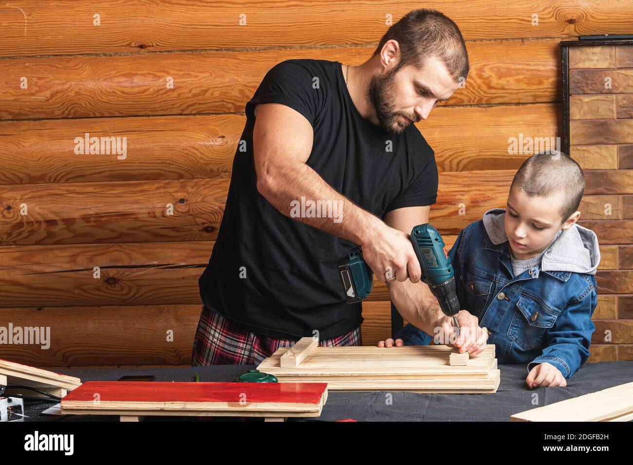 Papá e hijo están perforando una tabla de madera con un destornillador,  herramientas y una viga en la mesa en el taller. Concepto de entrenamiento  de carpintería para niños. Casa re Fotografía