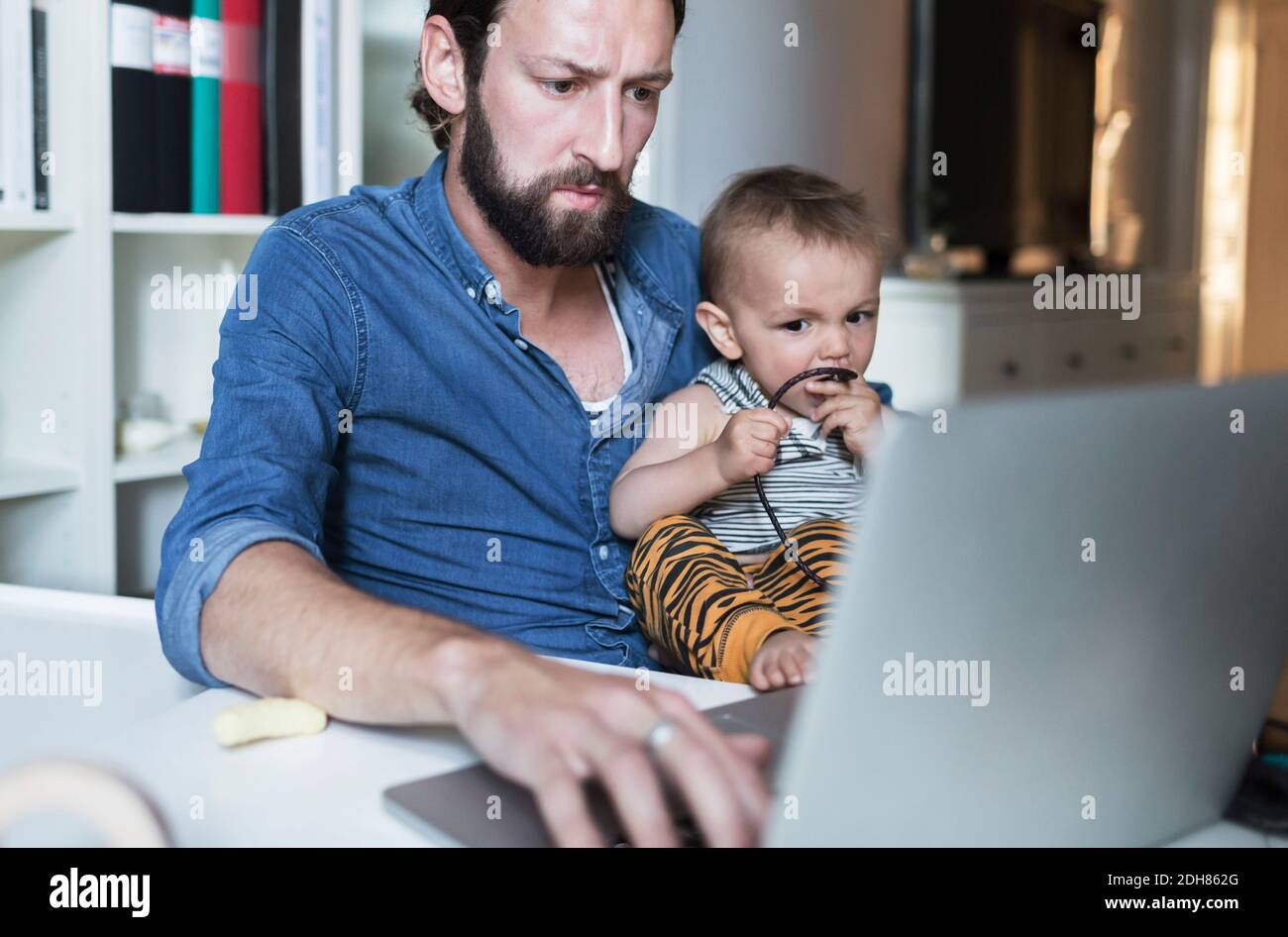 Hombre de negocios de adulto medio usando el portátil mientras cuidaba al bebé en casa Foto de stock