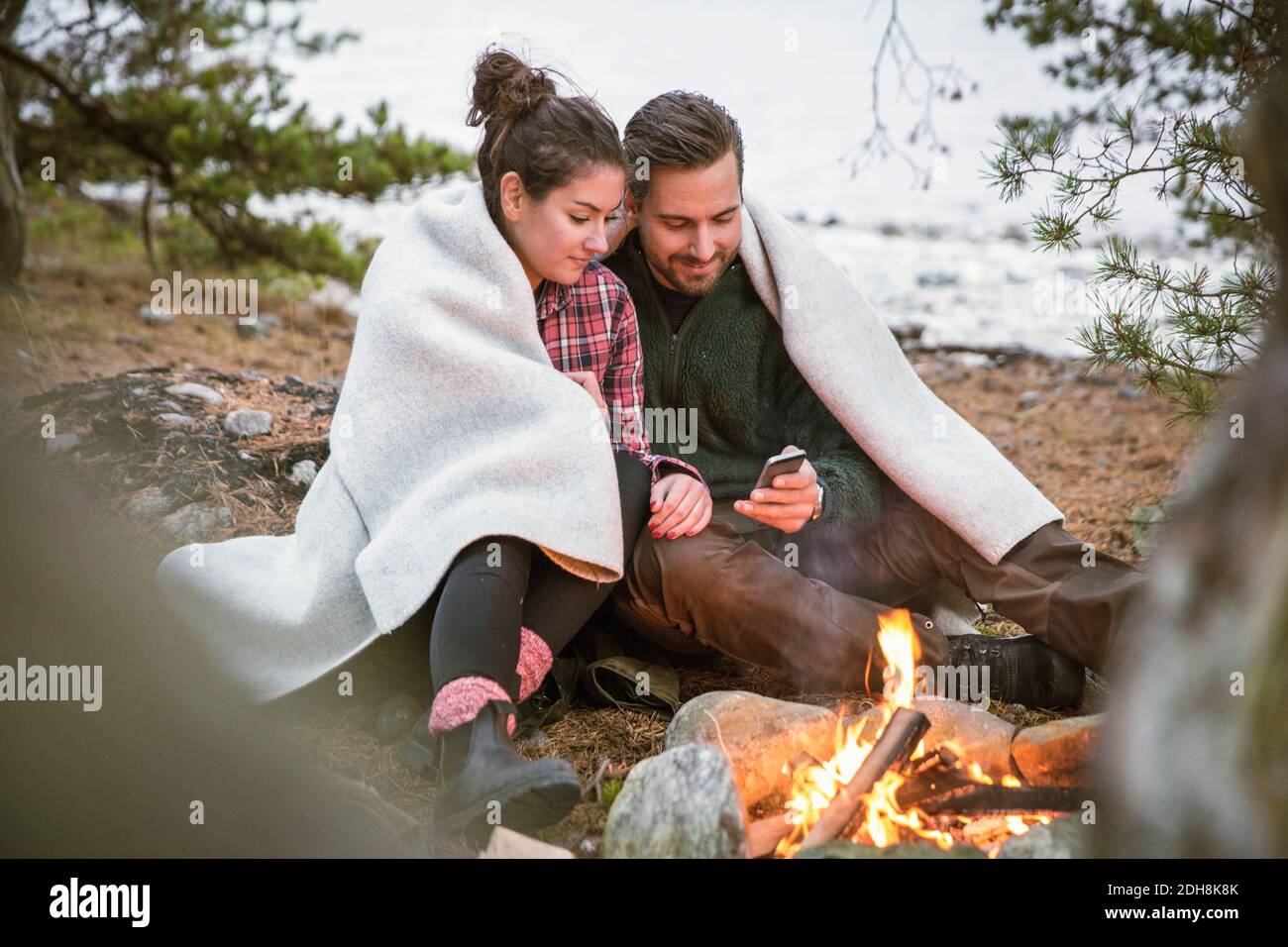 Una pareja usando el teléfono móvil mientras está envuelto en una manta por el fuego hoyo en el camping Foto de stock