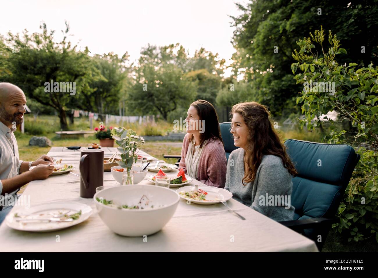 Mujer e hija sonriendo hablando con el padre mientras se sentaba mesa de comedor en el patio Foto de stock