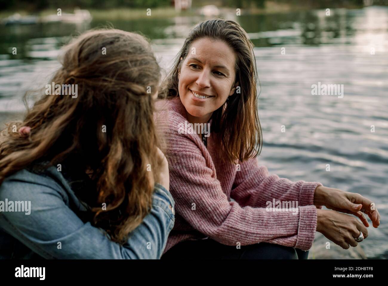 Madre sonriente hablando con su hija mientras está sentado en el lago Foto de stock
