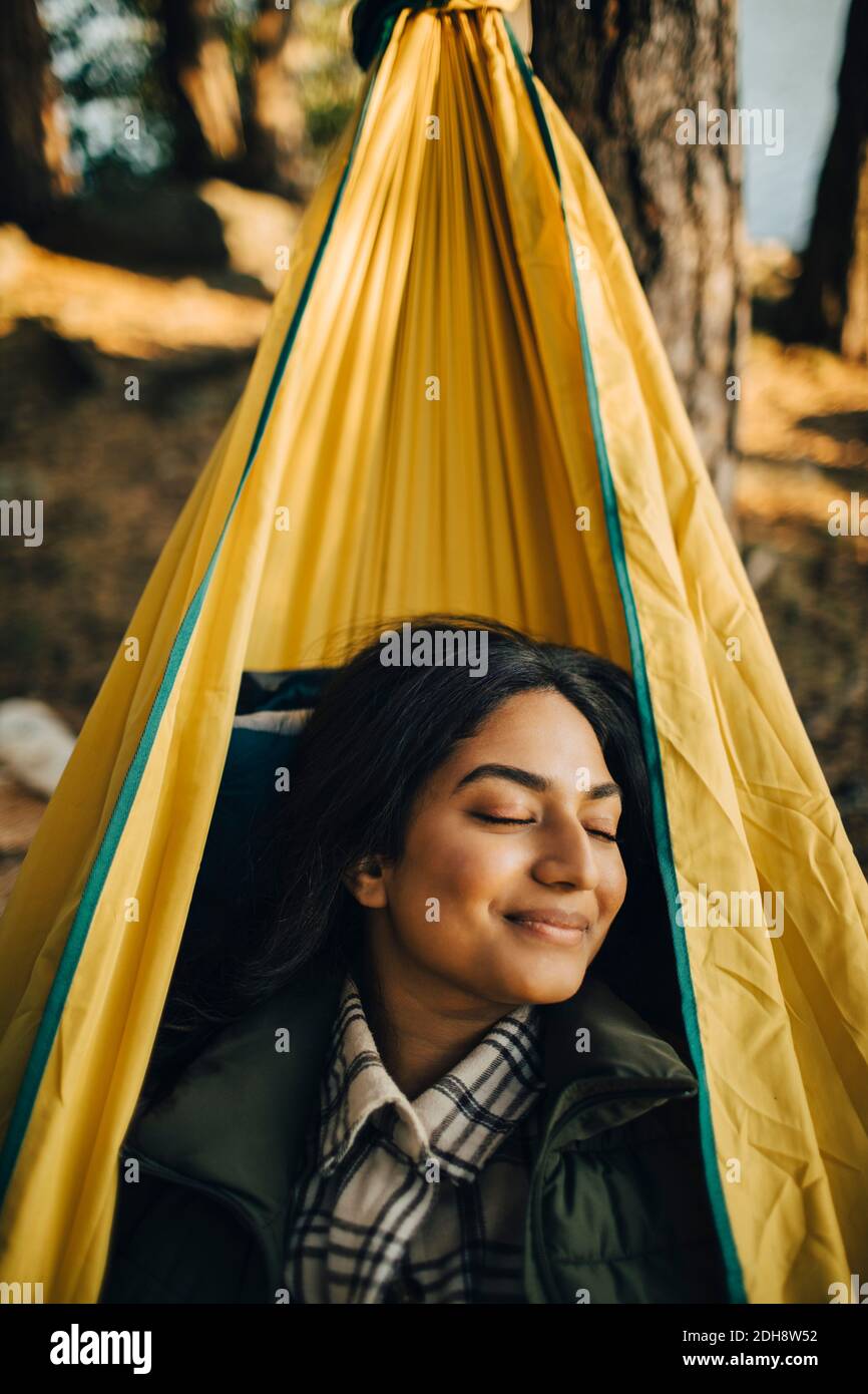 Mujer sonriente con los ojos cerrados en la hamaca en el bosque durante vacaciones Foto de stock