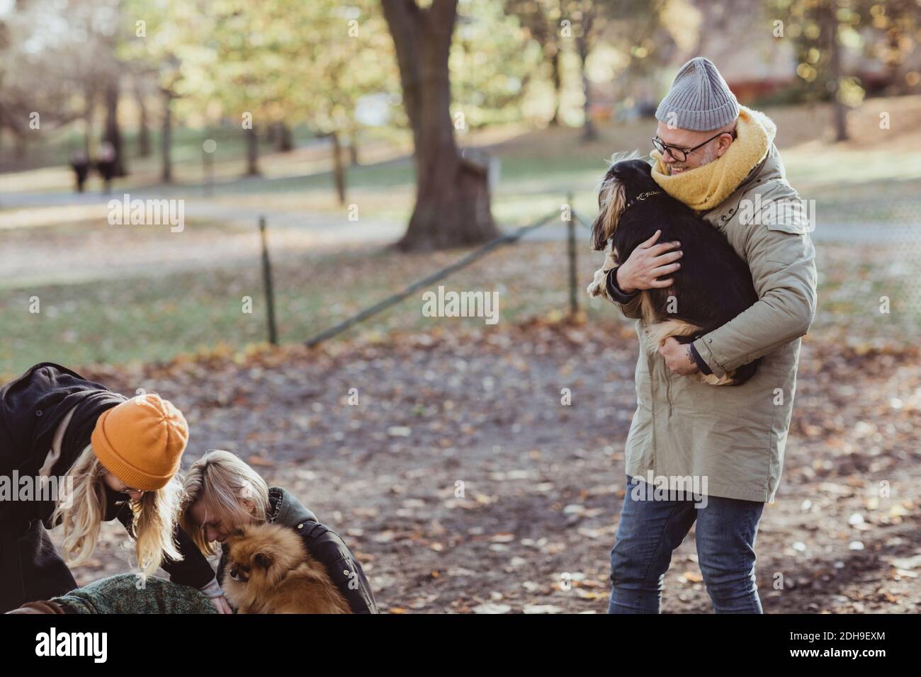 Hombre sonriente llevando perro mientras caminaba hacia mujeres y mascotas en el parque Foto de stock