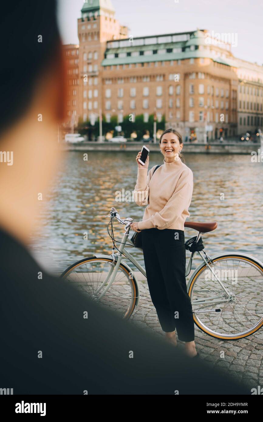 Mujer sonriente con bicicleta mirando a un amigo mientras está de pie río en la ciudad Foto de stock
