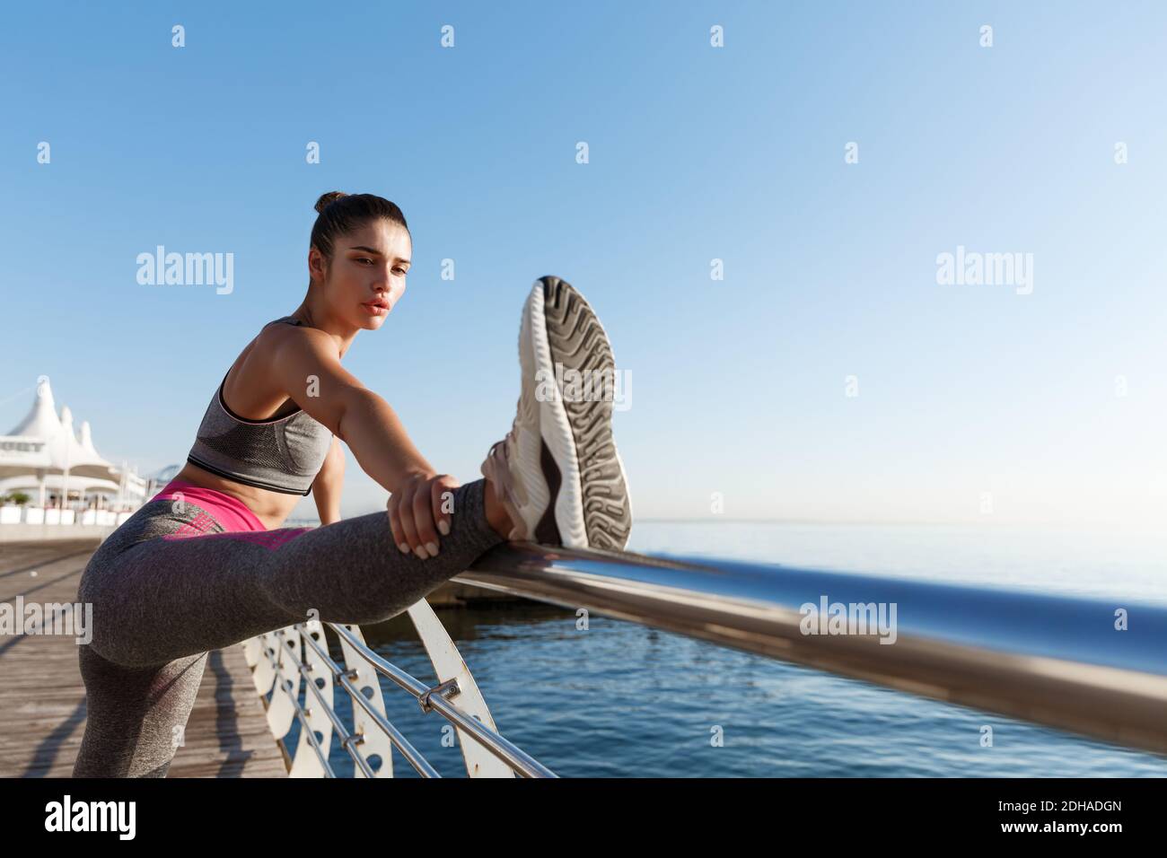 Tiro al aire libre de mujer atractiva y saludable con ropa deportiva, estirando las piernas y calentándose antes de correr cerca del mar Foto de stock