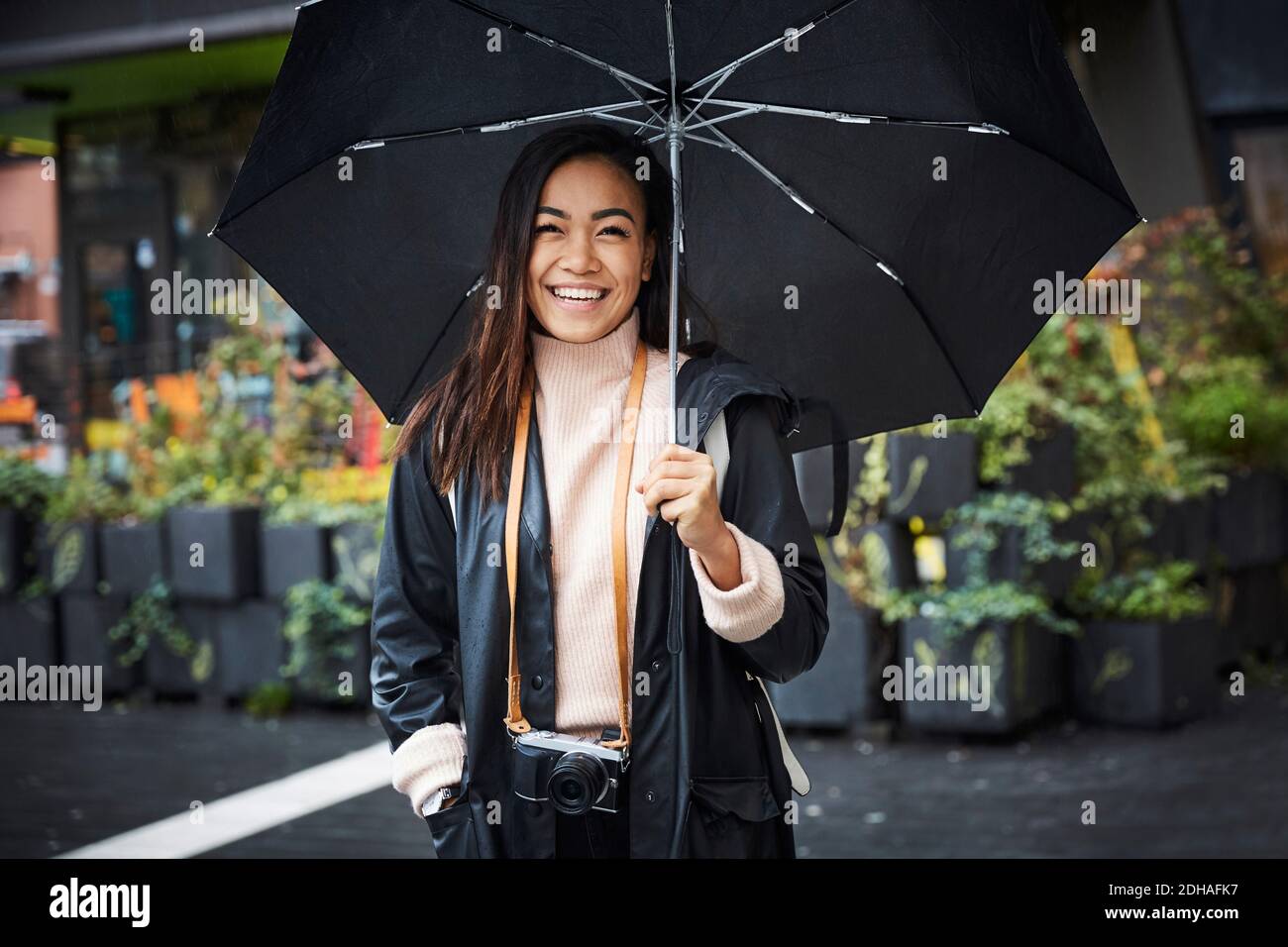 Mujer sonriente con cámara que lleva paraguas mientras está de pie en la ciudad Foto de stock