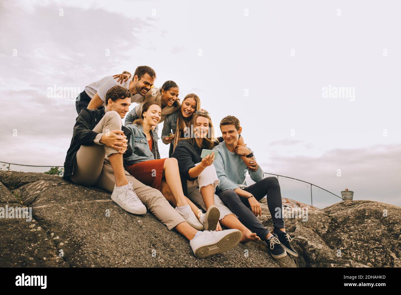 Feliz hombre y mujer amigos mirando el teléfono móvil mientras sentado en la formación rocosa contra el cielo Foto de stock