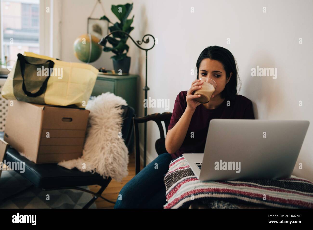 Mujer bebiendo té mientras usa el portátil en la pared durante la reubicación de casa Foto de stock