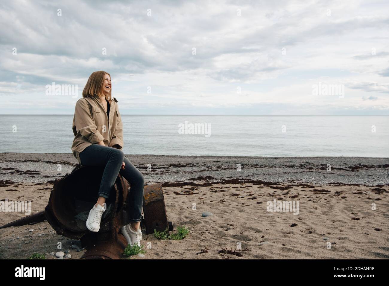 Alegre mujer joven sentada en el metal desgastado en la playa contra cielo Foto de stock