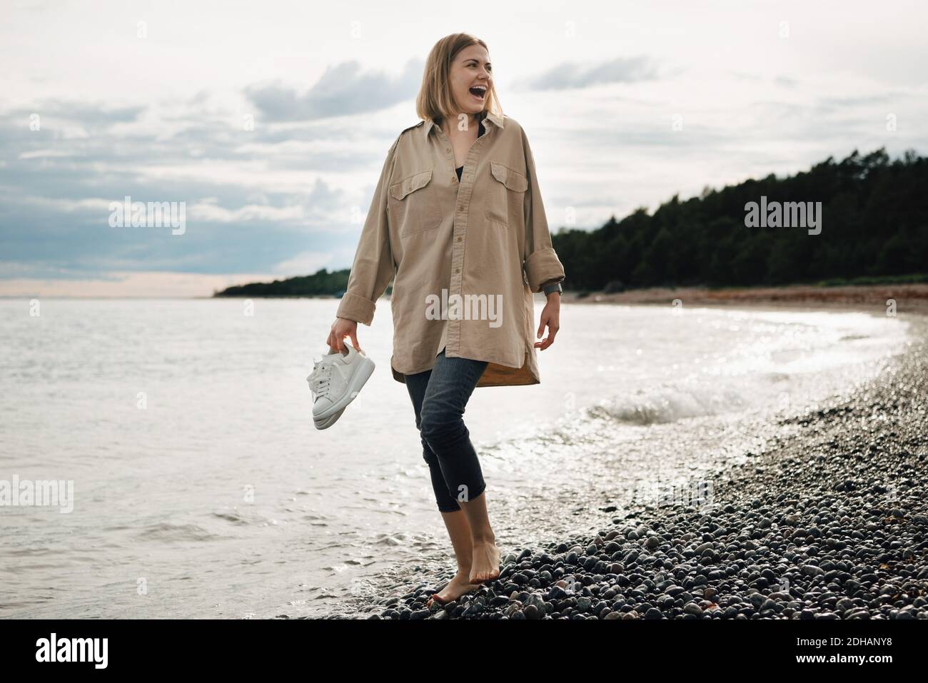 Alegre mujer joven sosteniendo zapatos mientras caminaba en la playa contra cielo Foto de stock