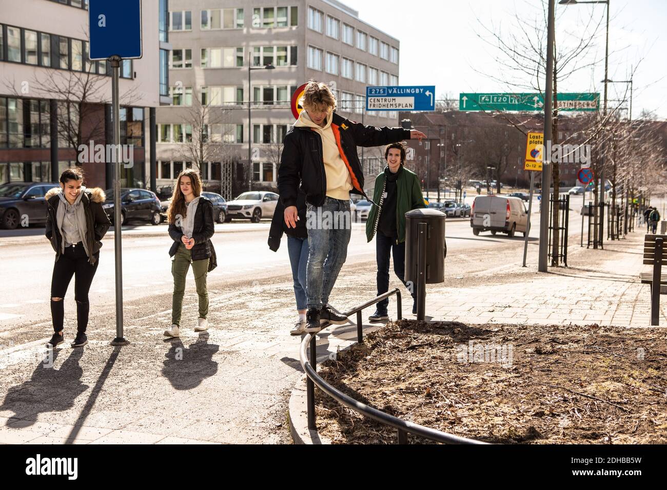 Adolescente balanceándose en la barandilla mientras los amigos caminan en la acera en la ciudad durante el día soleado Foto de stock