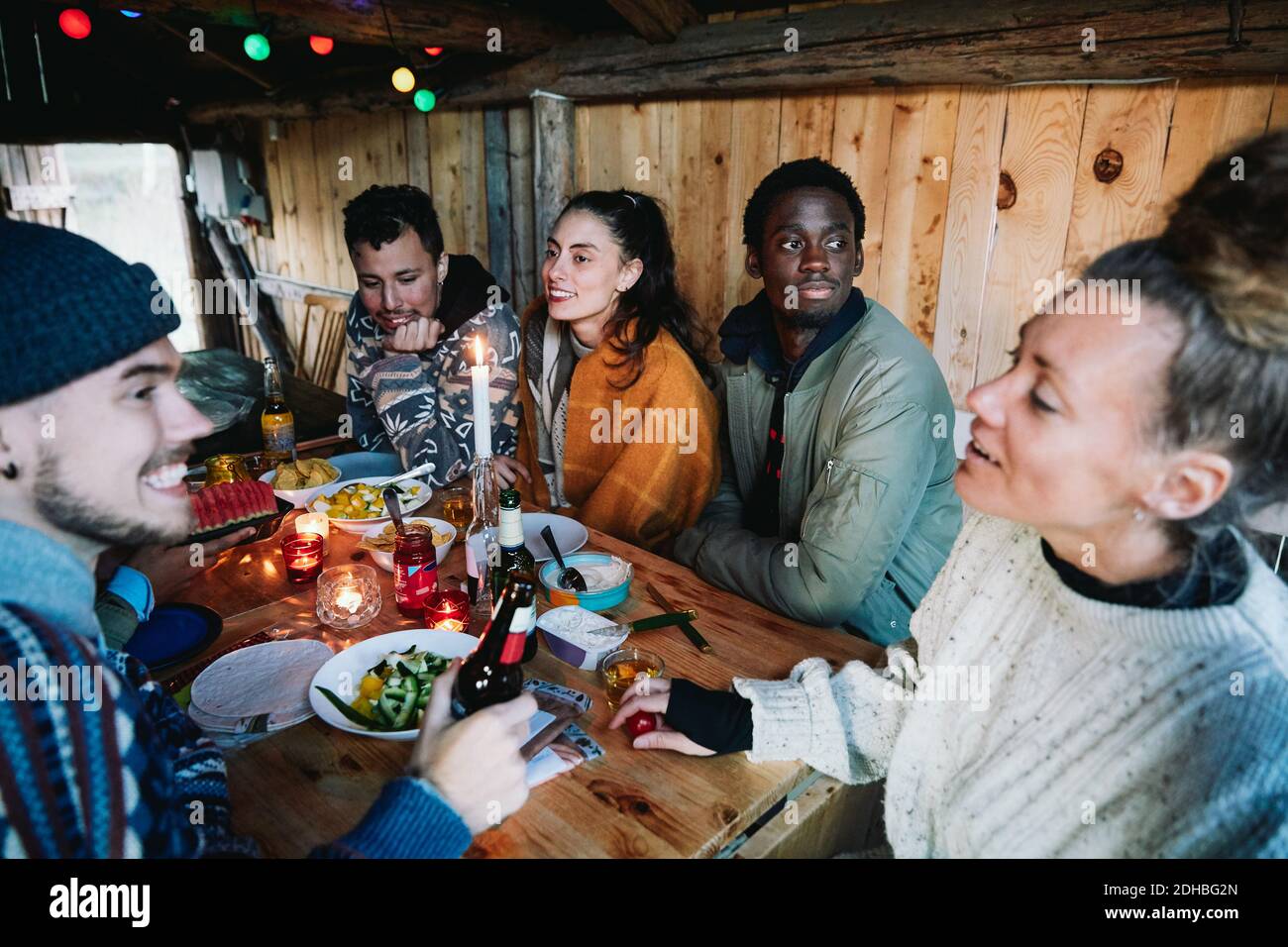 Amigos masculinos y femeninos hablando mientras tienen comida en la mesa en la cabaña de madera Foto de stock