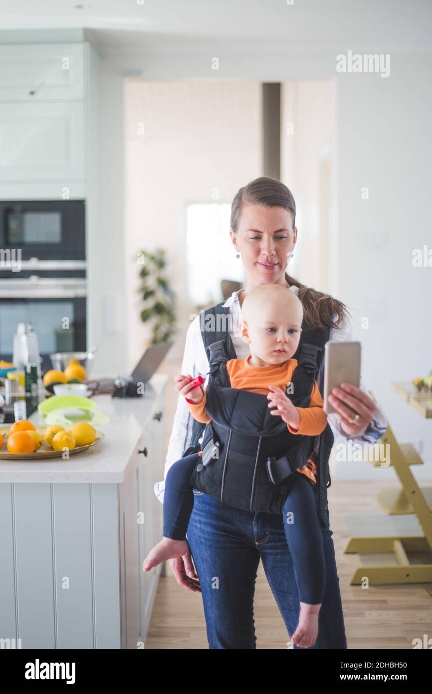 Mujer freelancer blogging mientras que lleva a hija en la cocina utilizando inteligente teléfono en casa Foto de stock