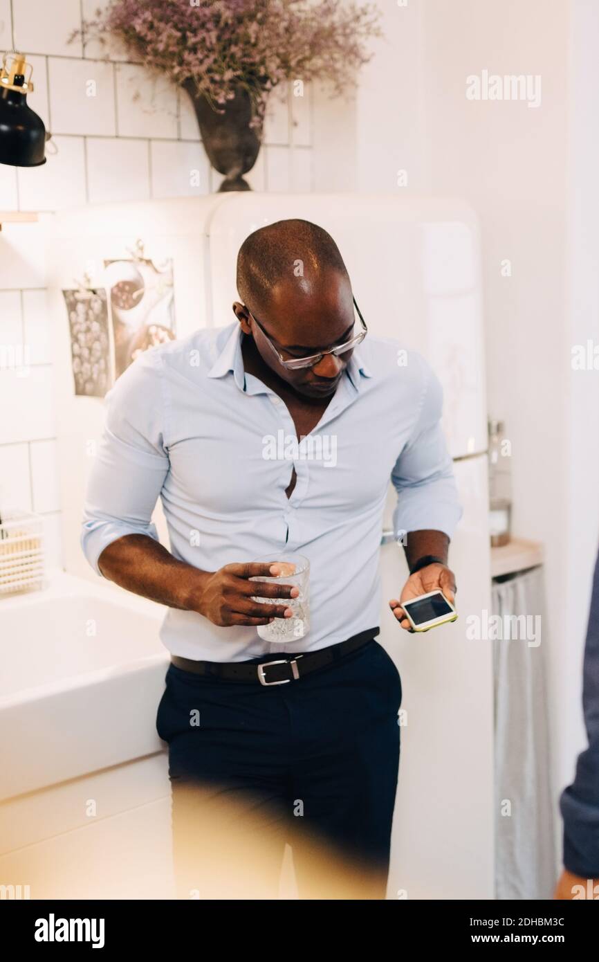 Hombre maduro usando el teléfono móvil mientras sostiene el vaso de beber adentro cocina en casa Foto de stock