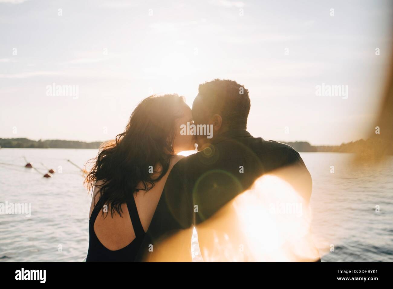 Pareja multiétnica besándose mientras se sienta contra el lago durante el verano Foto de stock
