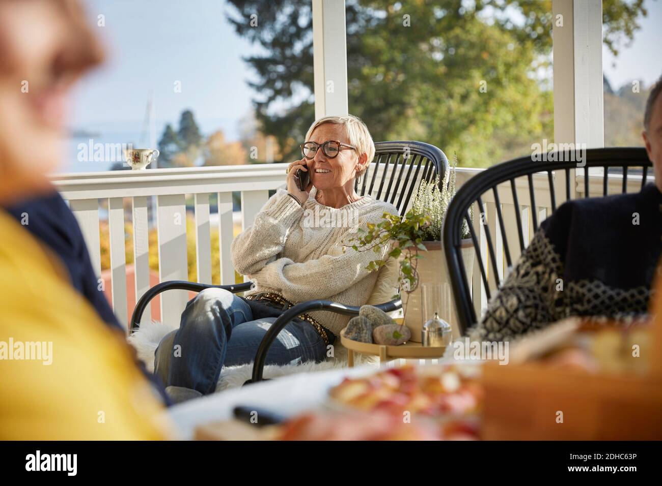 Sonriente mujer madura contestando teléfono inteligente mientras se sienta en el porche Foto de stock