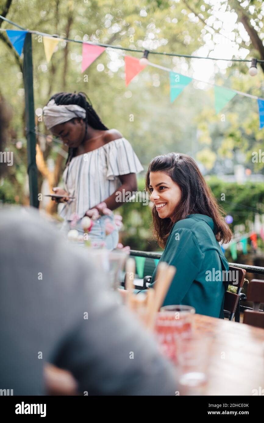 Una joven sonriente mirando hacia fuera mientras estaba sentada en una silla mesa en el balcón durante la cena Foto de stock