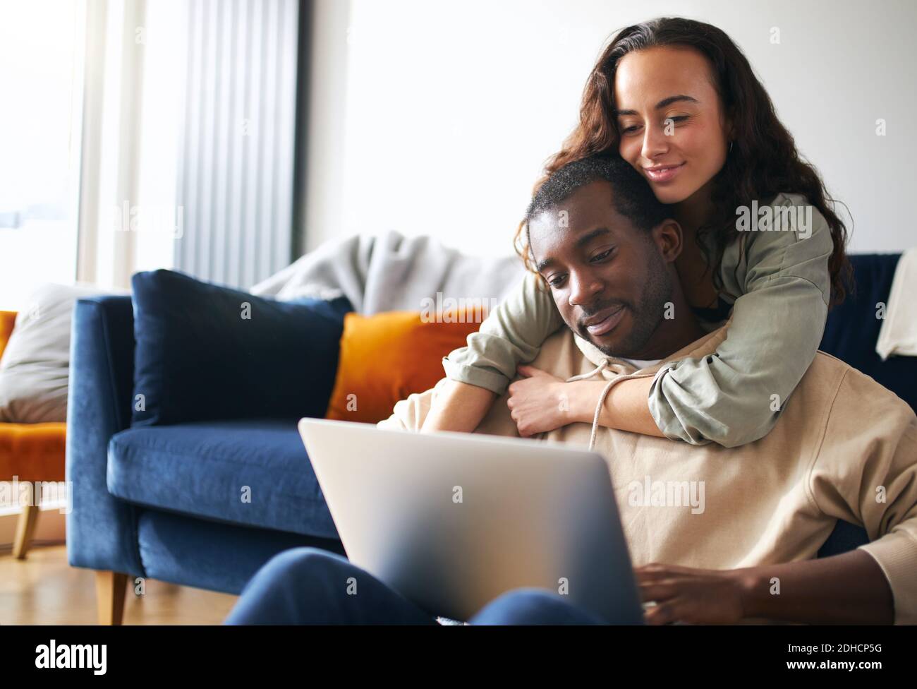 Mujer abrazando al hombre desde atrás como joven pareja de etnicidad mixta relájese en el sofá en casa navegando por internet en el ordenador portátil Foto de stock