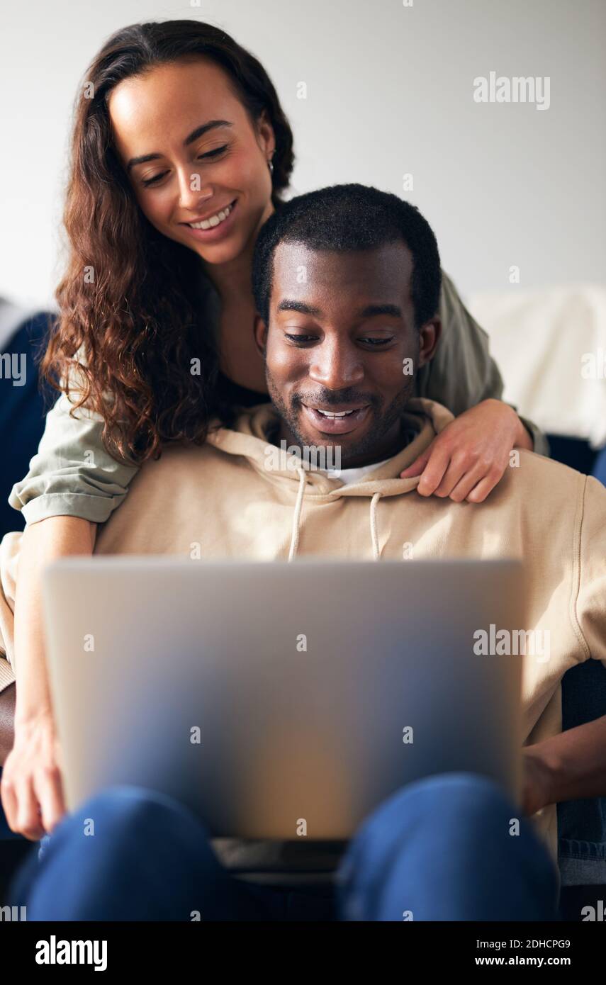 Mujer abrazando al hombre desde atrás como joven pareja de etnicidad mixta relájese en el sofá en casa navegando por internet en el ordenador portátil Foto de stock