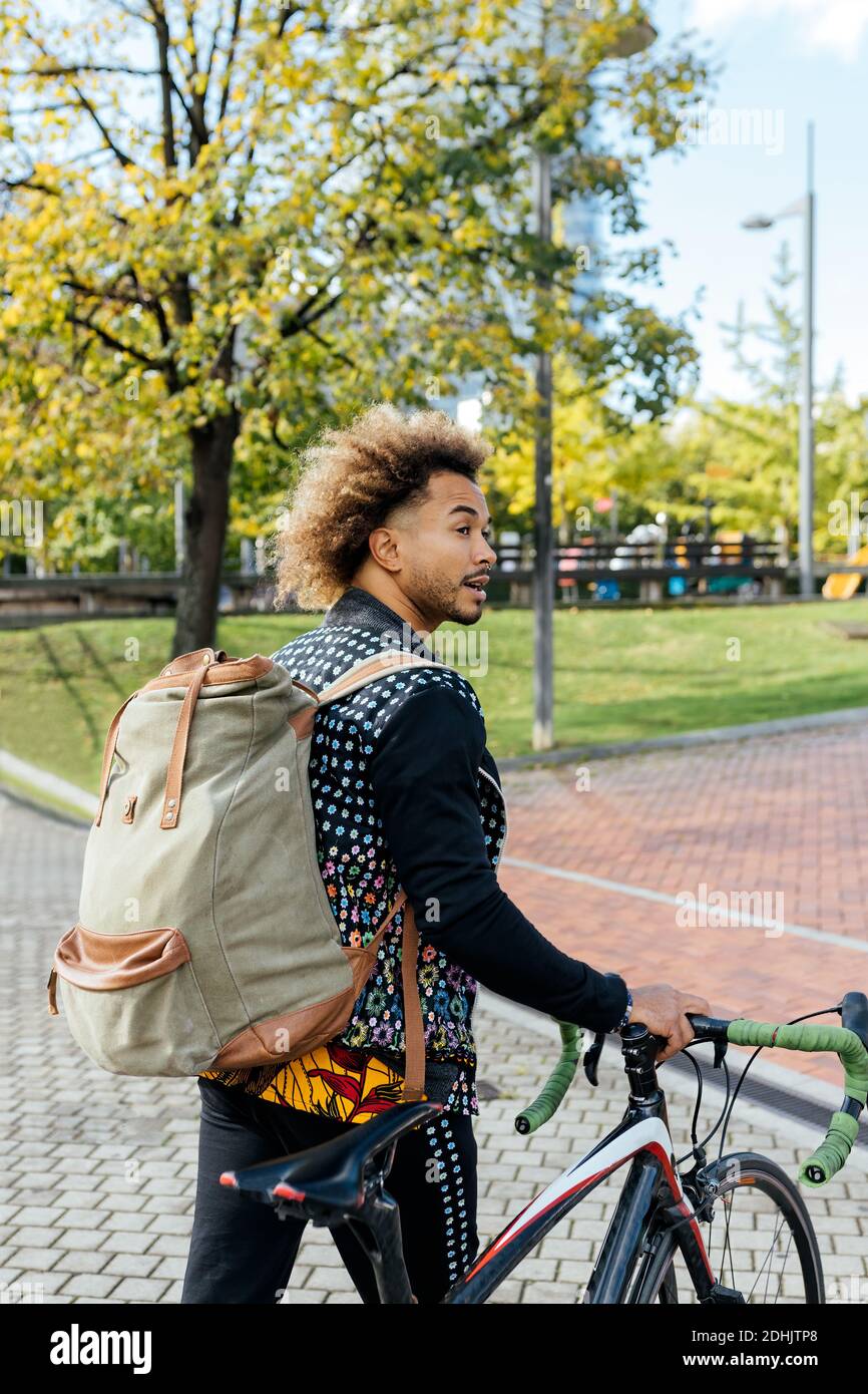Joven hipster de pelo rizado afroamericano masculino en moda con mochila de  pie con bicicleta en la plaza de la ciudad contra la moderna edificio alto  y looki Fotografía de stock -