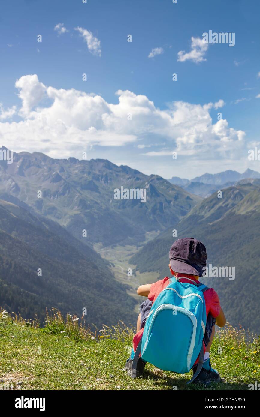 un niño aventurero e intrépido con un capuchón azul y una mochila, en cuclillas en la espalda, observa desde la cima de una montaña el valle de una montaña Foto de stock