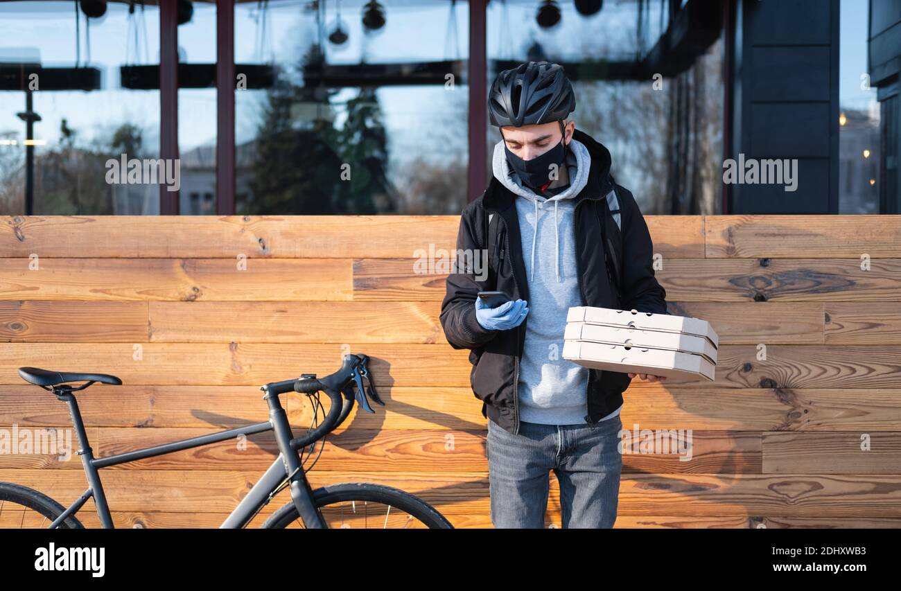 El hombre de la entrega de bicicletas en el casco sostiene un teléfono y  cajas de pizza. Entrega de comida, comida para llevar - bicicleta persona  de mensajero en el trabajo al