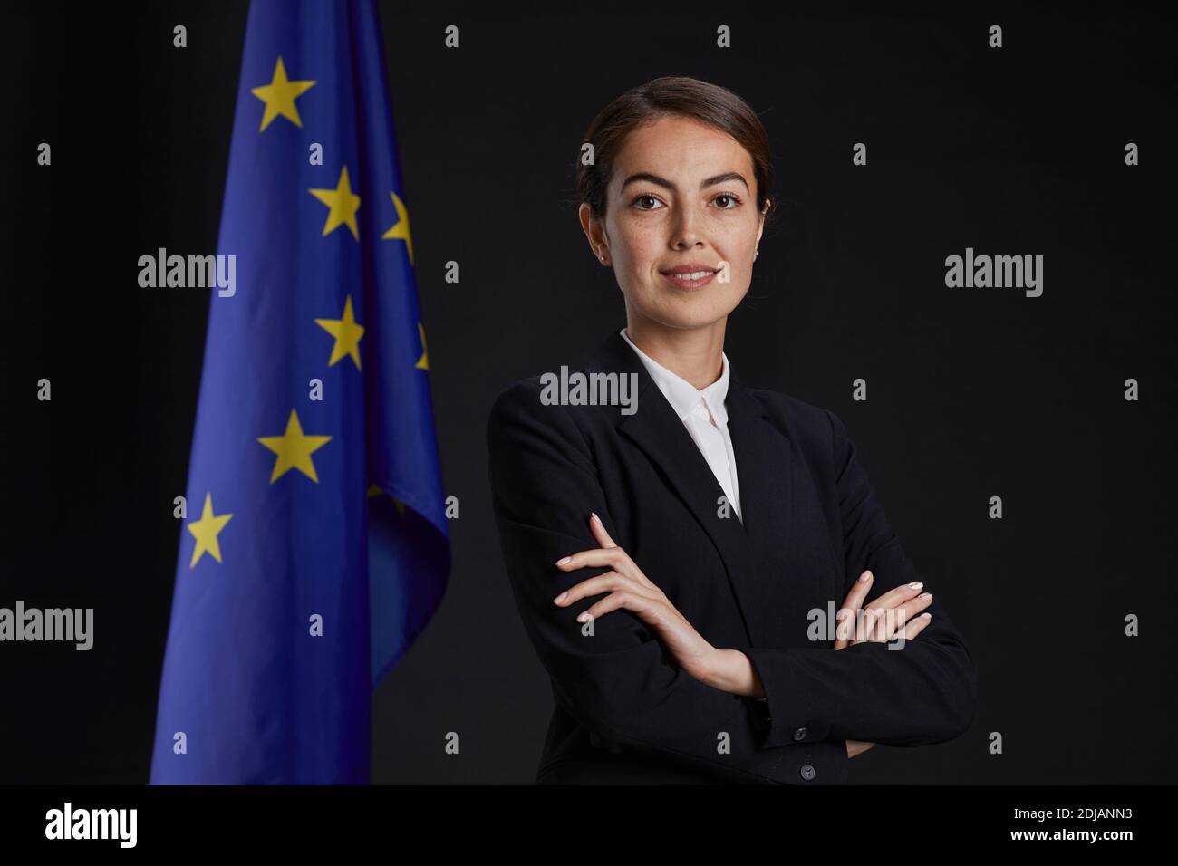 Retrato profesional de cintura arriba de una joven ejecutiva mirando la cámara y sonriendo mientras se pone de pie con los brazos cruzados contra la bandera de la UE en el fondo, Foto de stock