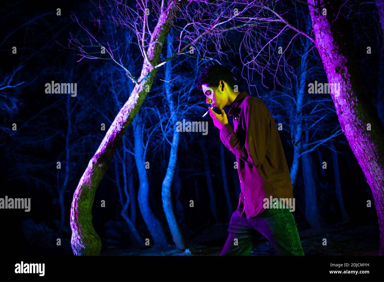 Vista lateral del Hombre Fumando Cigarrillo mientras camina por árboles iluminados en el bosque por la noche Foto de stock