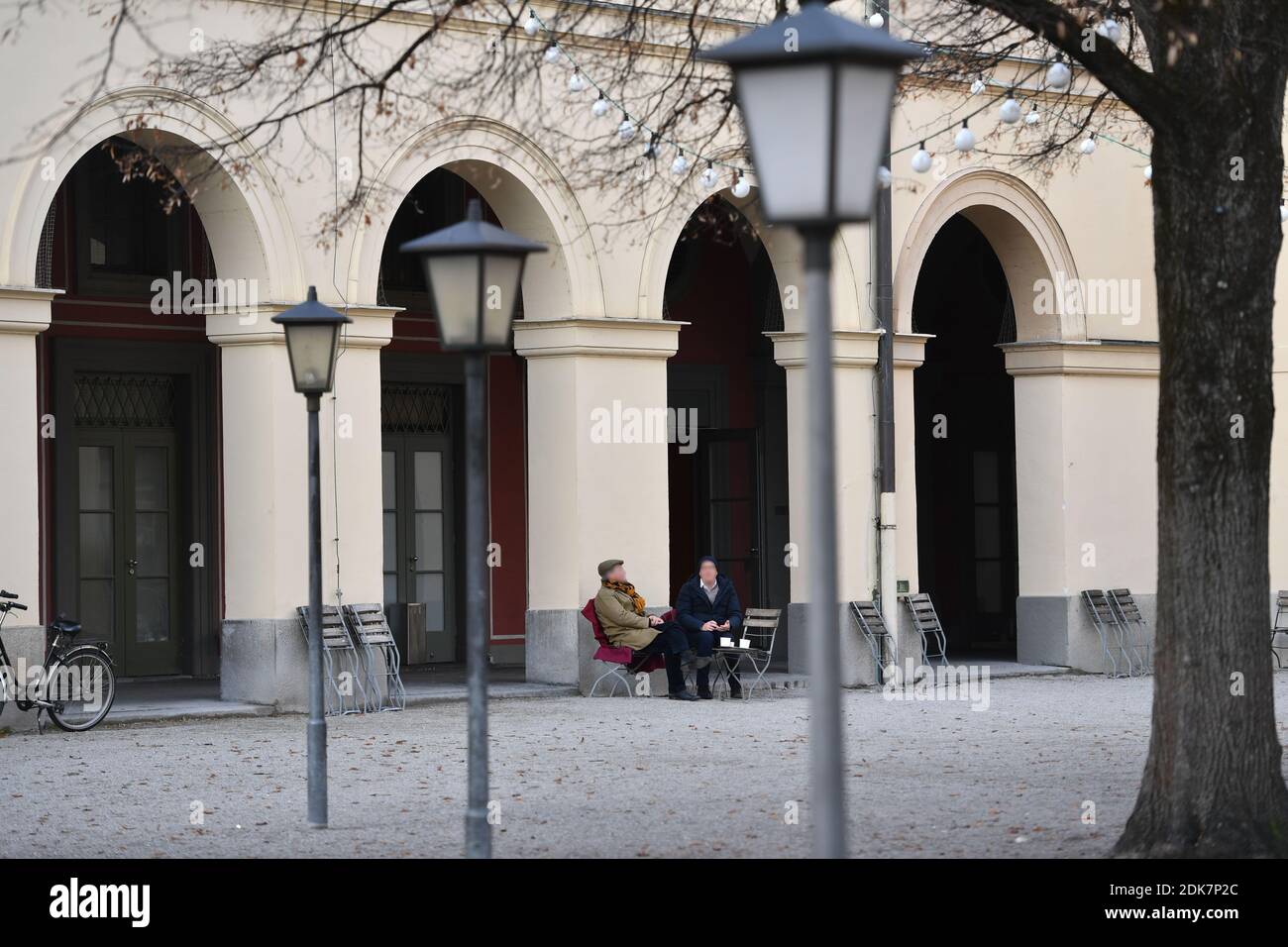 Foto del tema: Pandemia de coronavirus/consecuencias para la gastronomía. Dos transeúntes, la gente se sienta al lado de las sillas dobladas y bebe su café para ir. Mesas y sillas juntas en la zona exterior de un restaurante en el Hofgarten en Munich el 14 de diciembre de 2020. A partir del 16 de diciembre de 2020, se iniciará un cierre de emergencia hasta el 10 de enero, la restauración debe permanecer cerrada, consecuencia del cierre de la pandemia de corona, bloqueo, cierre, valor de incidencia. Uso en todo el mundo Foto de stock