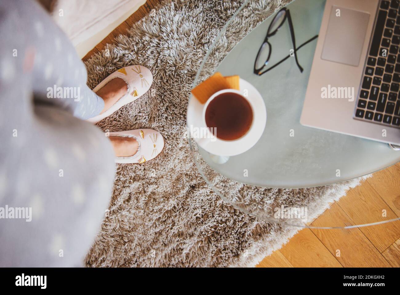 Los pies y las piernas de una mujer joven con ropa informal ropa de casa o pijama y zapatillas de gatito rosa y de pie en la alfombra cerca de la mesa de café con café Foto de stock
